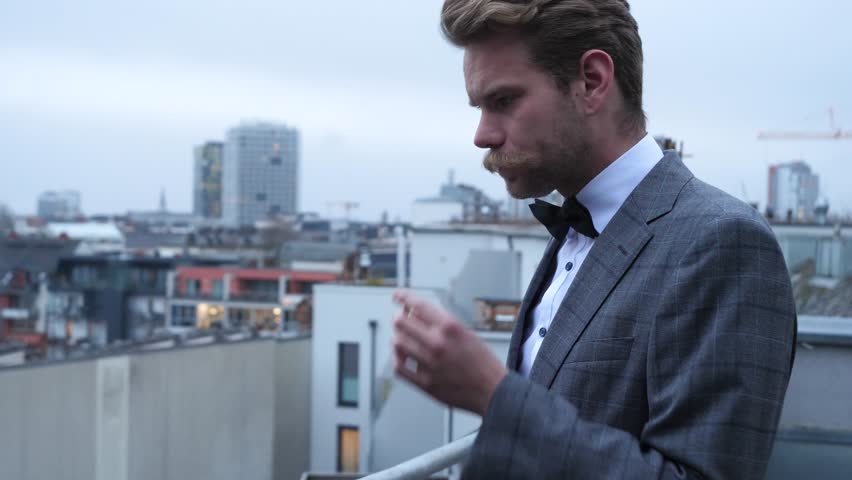 a gently suited man with blonde brown hair and a lovely mustache, smoking and looking away on a cold and foggy wintersday. In the backround is a city with small buildings and some crans.