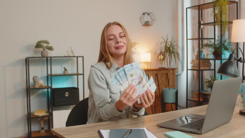 Businesswoman holding fan of cash money Ukrainian hryvnias UAH banknotes celebrate dance success career lottery jackpot game winner big income, wealth at home office workplace. Remote distant working.
