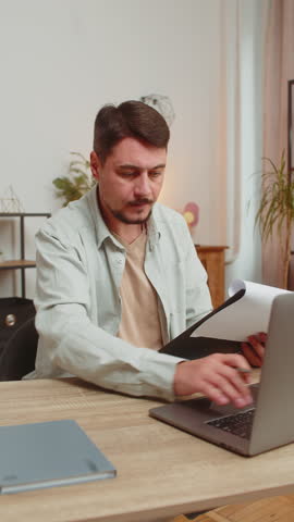 Caucasian freelancer accountant working with financial graph documents, calculate taxes for charts and reports at home office desk. Guy working on laptop computer, planning expenses budget. Vertical.