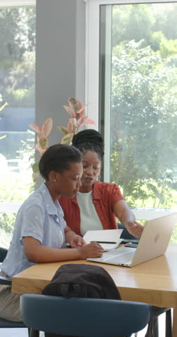 Vertical video: African woman pointing and guiding teen girl typing at laptop by window with notes. Mentorship, student, collaboration, guidance, learning, sunlit, vertical video