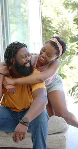 Vertical video: African American couple Youth woman coming playfully hugging Adult man on ottoman. Warmth, affection, laughter, embrace, sunlit, glassdoor, vertical video