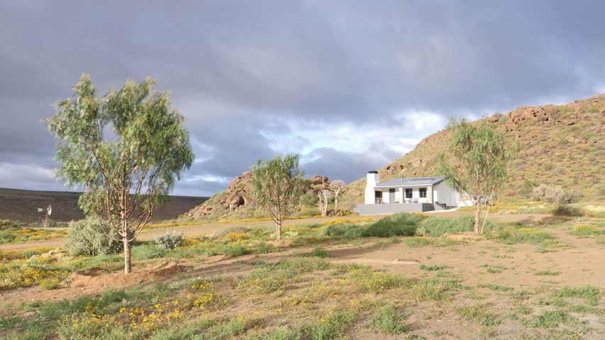 Remote guest farm in the Karoo with a white house, trees, and rocky hills under a cloudy sky