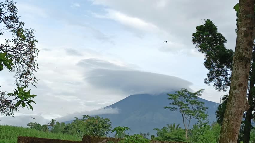 A breathtaking landscape of Mount Arjuno in Singosari, Malang, featuring a unique lenticular cloud formation at the peak while a flock of swiftlets (Burung Walet) flies gracefully across the sky during a cloudy morning.