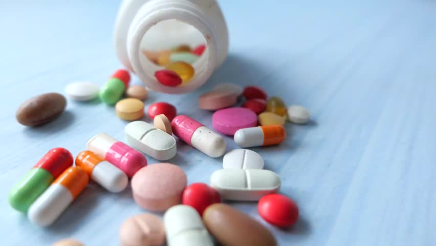 Close up shot of colorful pills and capsules spilling from an open white bottle onto a blue surface