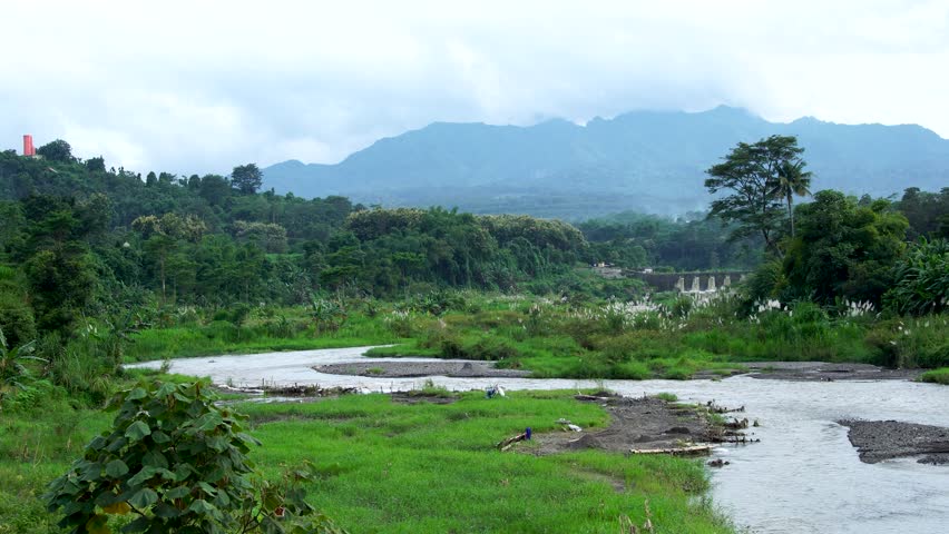 Panning shot of a winding river at a mountain base in Indonesia, Southeast Asia, showing sites of illegal private sand mining. Visualizes environmental exploitation and rural industry.
