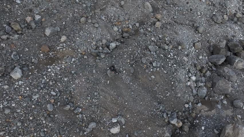 Aerial pull-out orbit shot revealing a man sitting on a jagged rock amidst a desolate, grey boulder field in Greenland.