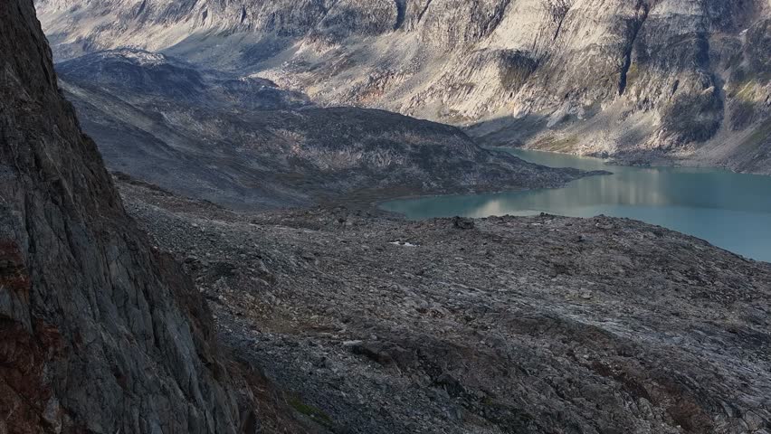 Majestic drone reveal around a vertical granite cliff wall in the remote mountains of Greenland overlooking a turquoise glacial lake.