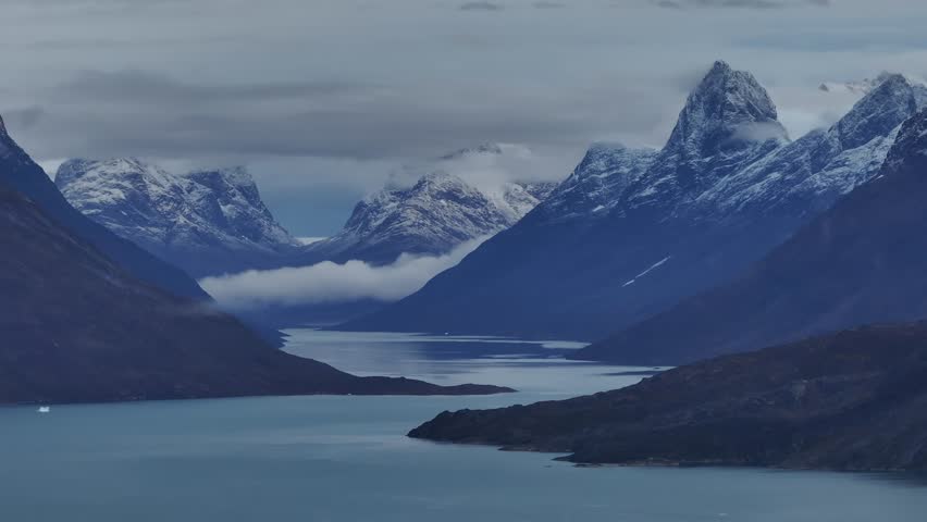 A slow aerial pan across the majestic snow-capped peaks and deep glacial fjords of Greenland under a moody overcast sky.
