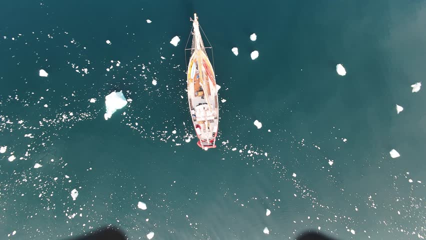Tracking aerial view of a red sailing yacht navigating through icebergs in the deep blue waters of Greenland, serene expedition footage.