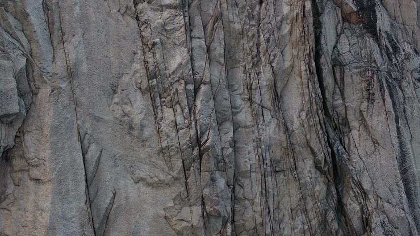 Cinematic reveal shot over a massive vertical granite cliff face revealing a turquoise glacial lake in Southern Greenland's desolate mountain wilderness.