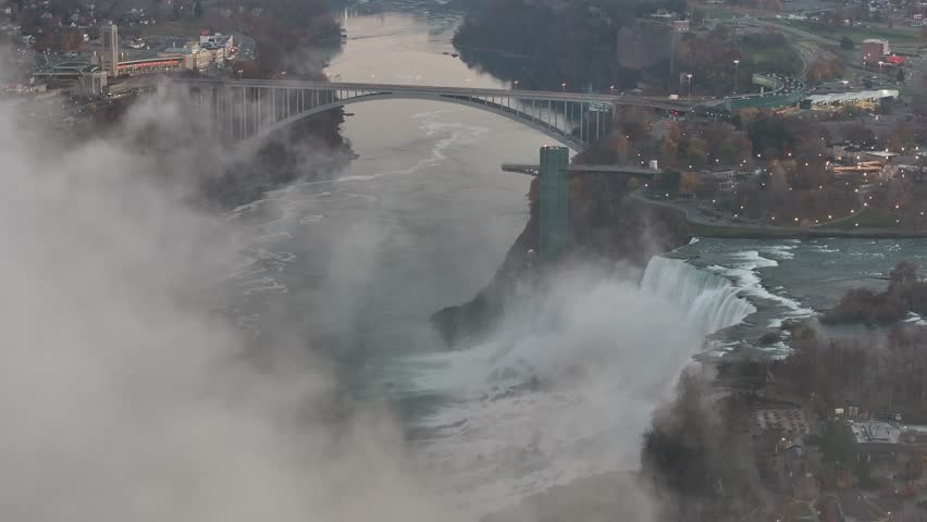 Majestic crane shot rising over the misty Niagara Falls and Rainbow Bridge at dusk, revealing the deep river gorge, 4K aerial footage.