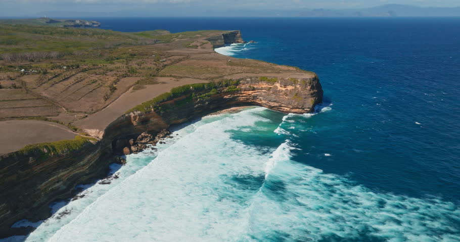 Indonesia rugged coastline features a dramatic panorama of steep orange cliffs meeting powerful turquoise ocean waves crashing onto the shore during a clear sunny day. Aerial view drone flight