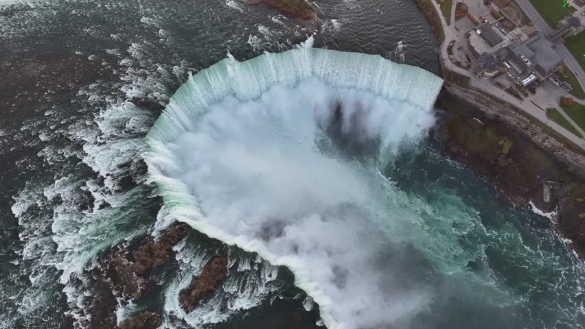 Majestic top-down aerial shot over the Horseshoe Falls in Niagara Falls, Canada, showing massive water volume and mist on an overcast day.
