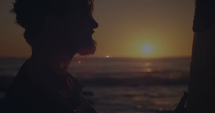 Man reaching up and lifting surfboard overhead while lens flare and ocean reflecting, prepping surf. Silhouette, sunset, beach, waves, horizon, shore, carrying