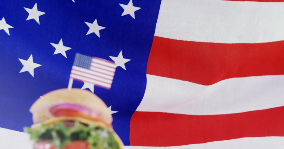 Camera pushing, stacked cheeseburger rising, mini US flag centering, bokeh drifting evoking pride. Sandwich, patriotic, studio, glow, lensflare, culinary, advertising