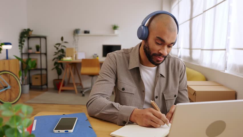 Young adult African American man taking notes during online course. Adult student learning from laptop while writing in notebook at home. Education and e-learning concept. 