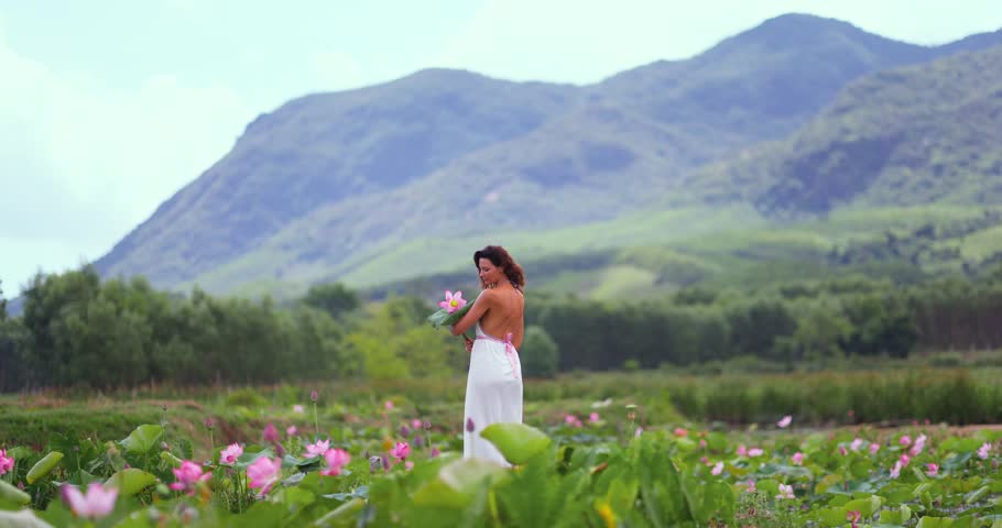 Cinematic woman in lotus meadow in Tra Ly, Duy Xuyen, Quang Nam, Vietnam, holding pink bloom against lush mountain backdrop. Scenic travel moment perfect for tourism campaigns, wellness visuals, and