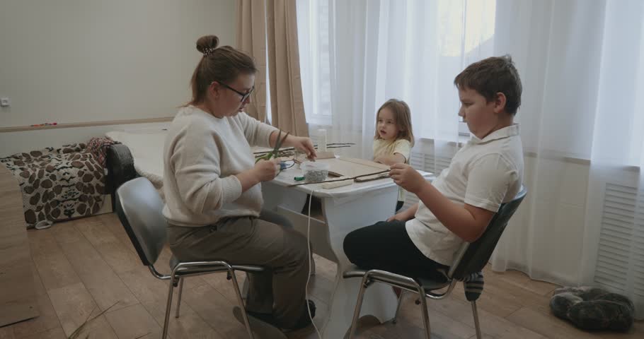 Loving family with mother, son, and daughter enjoying a creative crafting session together, sitting at a table in their living room and working with cardboard, scissors, and a hot glue gun.