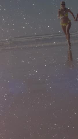 Vertical video: partner pulling woman running toward camera, beach marketing with sparkles wet sand. Runner, shoreline, ocean, reflection, glitter, bikini, vertical video