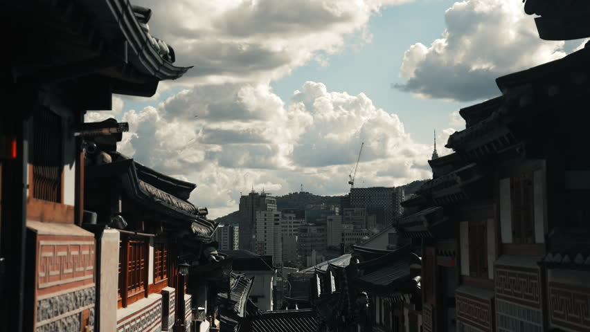 View through traditional Bukchon Hanok Village rooftops toward modern Seoul skyline and Namsan Tower under dramatic clouds