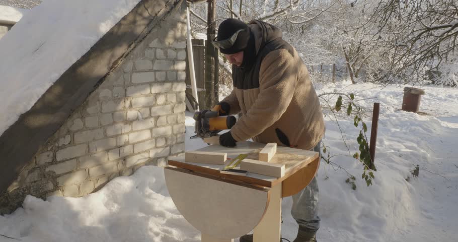 Young man using a handheld circular saw to cut a wooden plank for a bird feeder, working outdoors in a snowy backyard on a sunny winter day. Family project building a diy nesting box together.