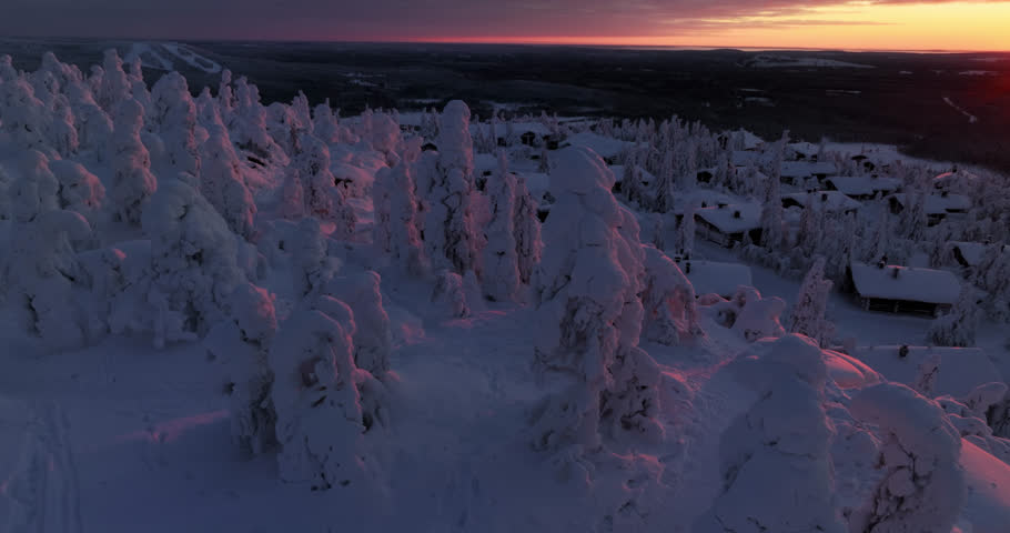 Aerial view around snowy trees, revealing mountain cottages, winter sunset in Lapland