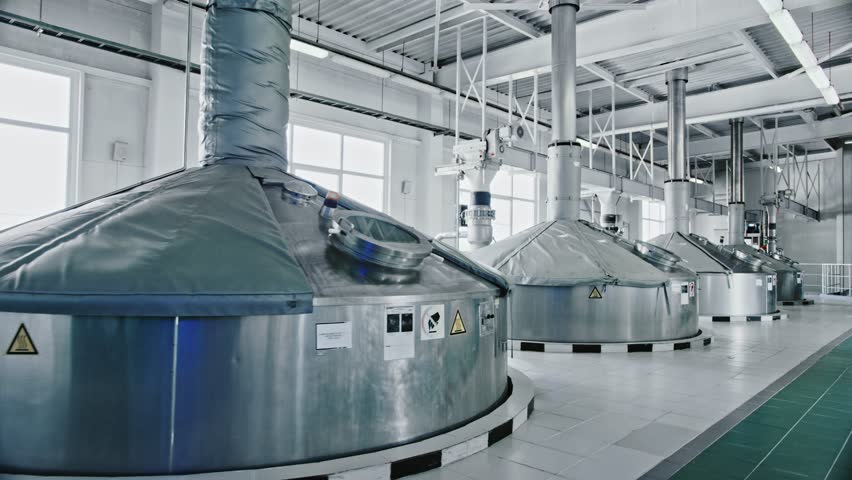 Large stainless steel vats and fermentation tanks standing in a row inside a clean, modern beer factory, showcasing the industrial equipment used for brewing and alcohol production process