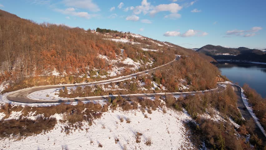 Aerial view of a car driving on a curvy road in a snowy mountain landscape next to a lake, showing a beautiful scenic drive during winter on a sunny day with blue sky and some clouds