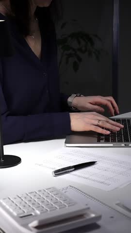 Woman working late on laptop, hands typing on keyboard, managing business paperwork at desk in office at night. Audit and taxes in business