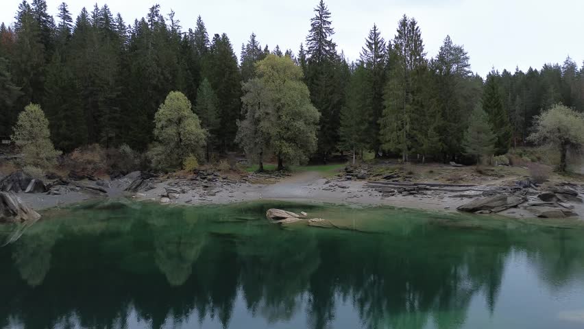 Aerial drone pull out revealing clear emerald lake surrounded by dense forest with reflections on calm water creating peaceful natural landscape scene.