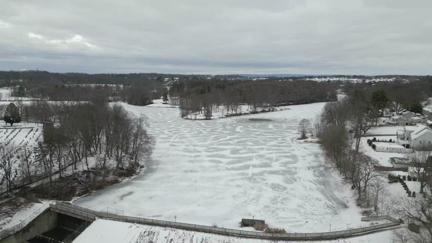 A cinematic high-angle drone shot capturing a wide frozen river surrounded by snow-covered trees and a white winter wilderness.