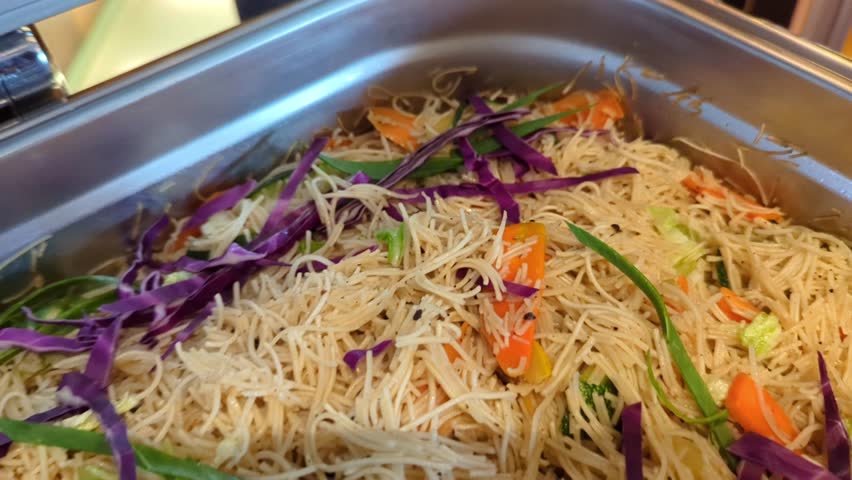 Close-up of freshly prepared vegetable noodle salad in a stainless steel catering tray, featuring thin noodles, cabbage, carrots and green onions in a vibrant buffet setting.