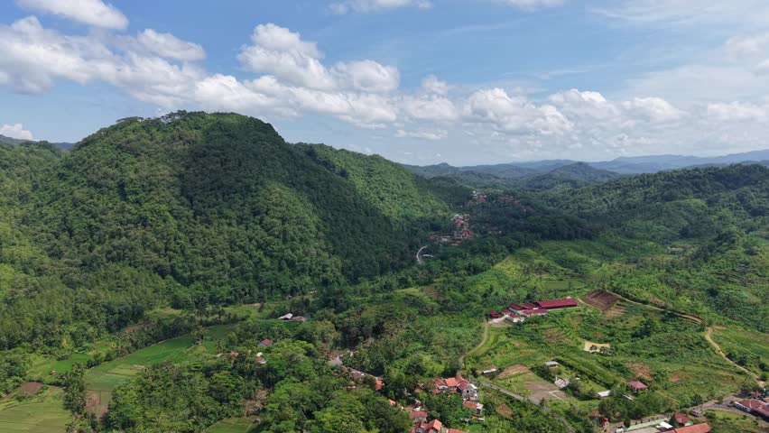 Lush green hills and a winding valley with a small village and farmland seen from above, showcasing tropical forested mountains, rural roads, and layered distant ridges under a bright sky.
