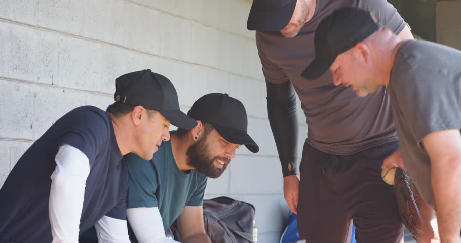 Showing smartphone prompting Diverse male teammates reviewing plays in dugout, with baseball glove. Athletics, teamwork, camaraderie, recreation, communication, leisure, outdoor