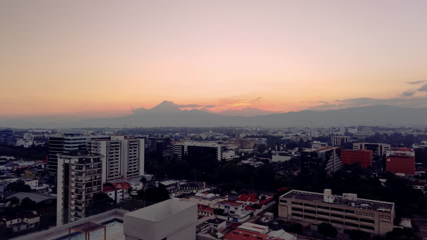 Slow drone approach over Guatemala City buildings during twilight. The cityscape is framed by a vibrant orange sunset and the distant silhouettes of Agua and Fuego volcanoes.