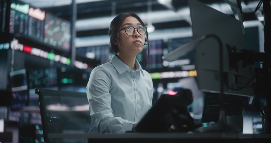 Female Trader Analyzing Real Time Stock Market Data at Large Screens. Focused Asian Woman in Glasses Works Intently at High Tech Desk, Typing on Keyboard While Monitoring Complex Financial Charts.