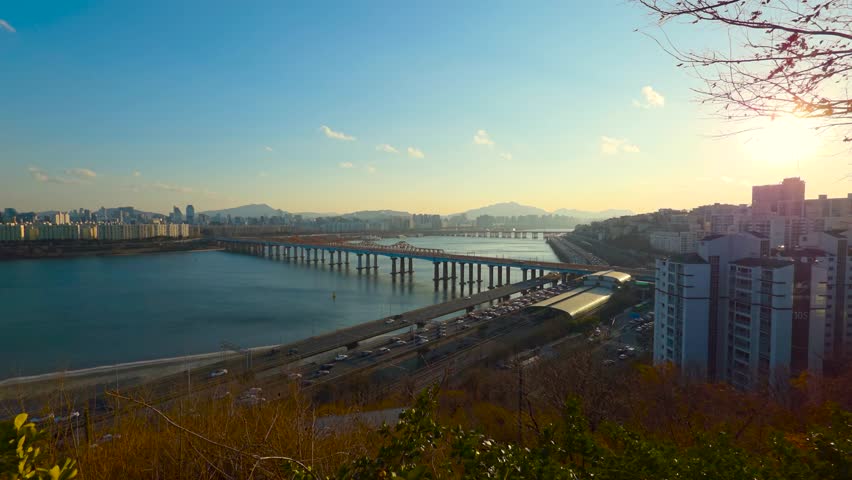 Real-time sunset zoom from hilltop revealing Donho Bridge, congested Gangbyeonbuk-ro expressway and glowing Oksu Station roof along Han River in Seoul against bright setting sun