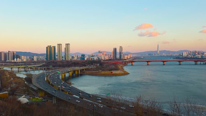 Real-time aerial of curved Gangbyeonbuk-ro expressway traffic at golden sunset with red Seongsu Bridge, Lotte Tower and Seoul skyline on Han River