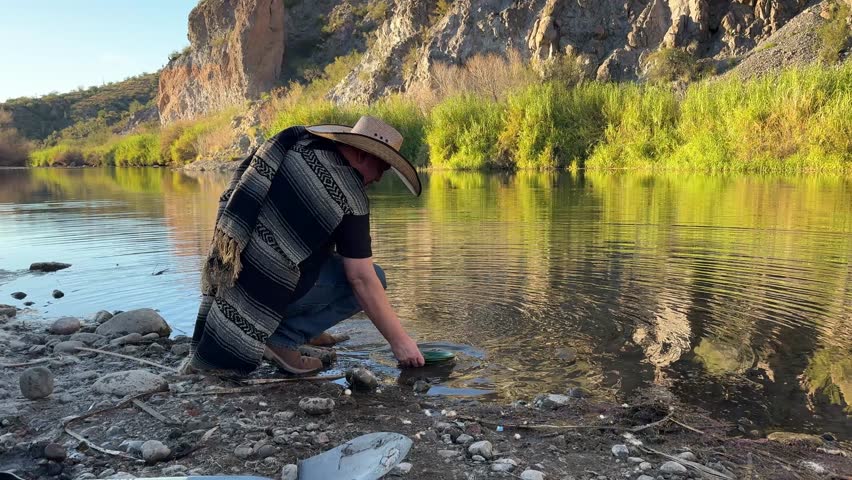 Millennial man (early 40s) wearing a poncho and cowboy hat, panning for gold in Arizona in river
