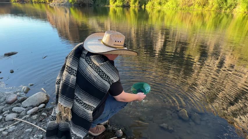 Millennial man (early 40s) wearing a poncho and cowboy hat, panning for gold in Arizona in river