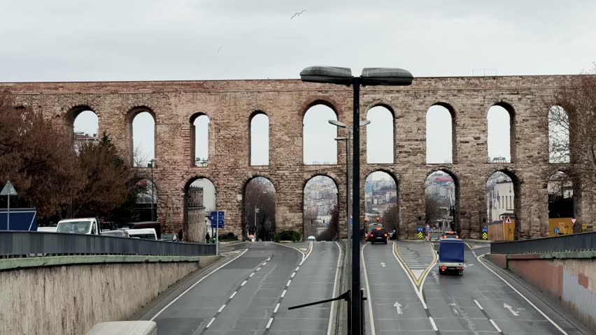 Historic Valens Aqueduct structure spanning divided road with vehicles moving beneath arches in urban environment during daytime, Istanbul, Turkey, Handheld, 4k