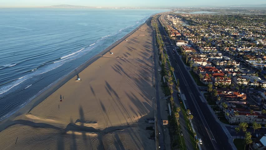 Huntington City Beach stretches west of 15th St with lifeguard towers lining shoreline near PCH frontage. Soft wave patterns, palm shadows, warm roof, coastal transition and tourism corridor, CA. USA