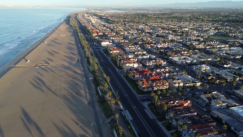 Coastal corridor along PCH parallels wide Huntington City Beach shoreline north of pier district near 15th St. Long palm shadows, clean sand bands, dense roof textures coastal urban divide, CA. USA