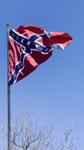Vertical video shows a large confederate flag flapping in slow motion in a stiff breeze on a flagpole in south Georgia.