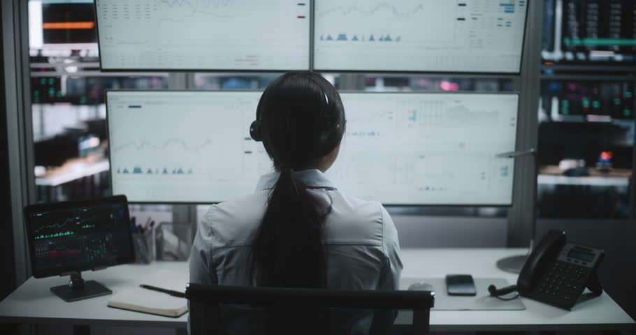 Trader in Headset Working at Sophisticated Workstation. Woman Interacts With Digital Tablet While Monitoring Four Large Screens Filled With Complex Stock Market Charts, Real Time Analytics. Back View.