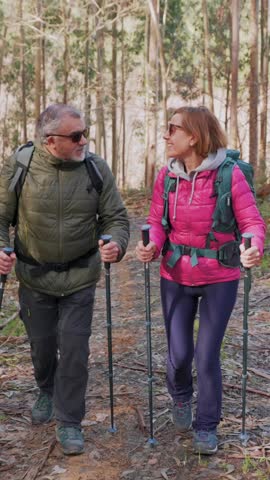 vertical Happy senior couple with backpacks and trekking poles hiking on a forest path, enjoying an active retirement lifestyle and the beautiful nature during their healthy walk together