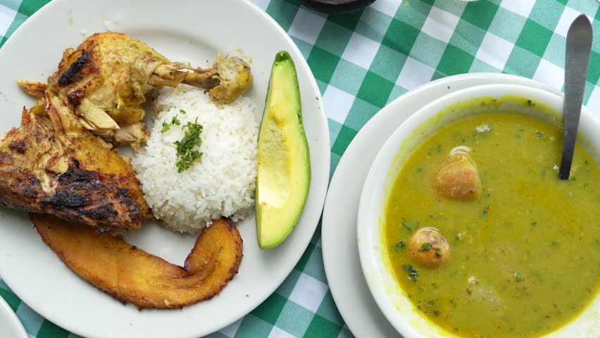 Close-up of delicious Colombian sancocho de gallina with rice, avocado, and fried plantains on a vibrant checkered tablecloth. Warm, inviting meal in soft natural lighting.