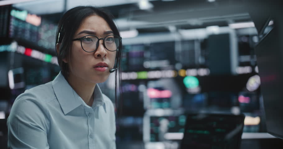 Focused Asian Businesswoman Operates at Multi Monitor Workstation in Modern Stock Exchange Hub. Focused Female Trader in Stylish Eyeglasses Reviewing Real Time Financial Data on Screens and Tablets.