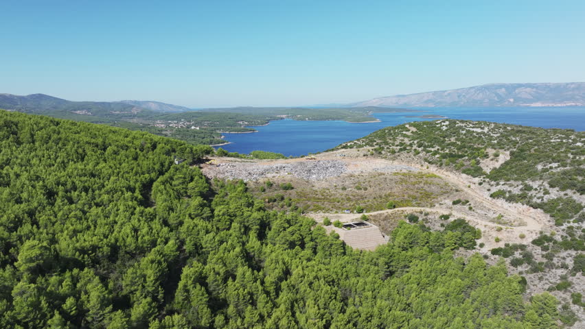 AERIAL: Expansive landscape of Hvar island with a waste management site surrounded by dense pine forests and scenic view of deep blue Adriatic Sea. Human impact and the natural coastal environment.