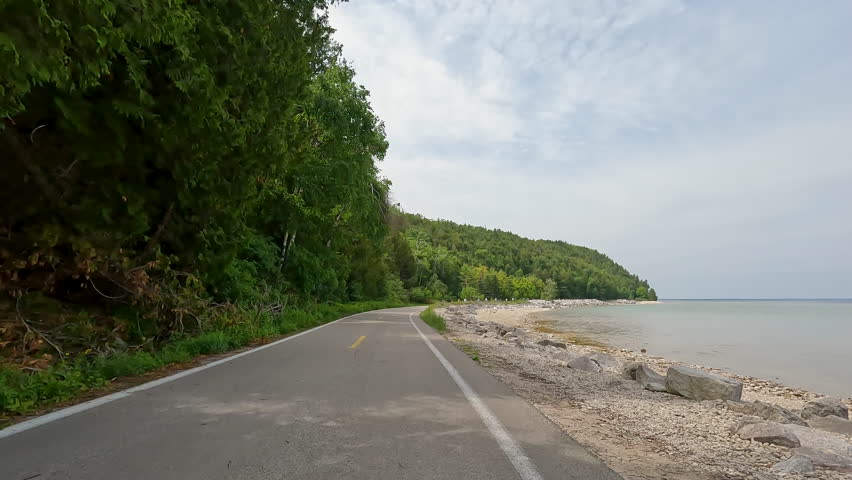 POV Biking on M-185 Highway Along Lake Huron Coast, Mackinac Island, Michigan, USA. POV slow-motion cycling on M-185, the car-free coastal highway on Mackinac Island, Michigan. The camera moves along the paved shoreline path of Lake Huron, featuring a rocky coast, turquoise water, and lush green forest under a bright sky.